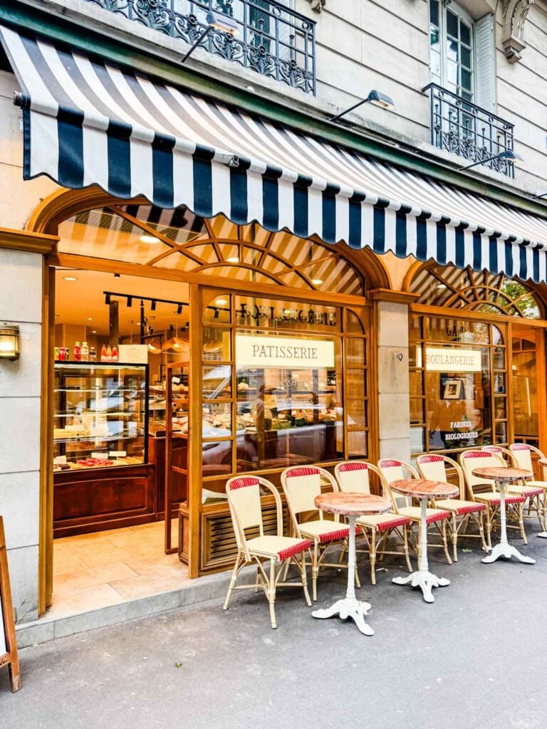 A blue and white awning hangs over cafe tables surround by chairs in front of local boulangerie in Paris.