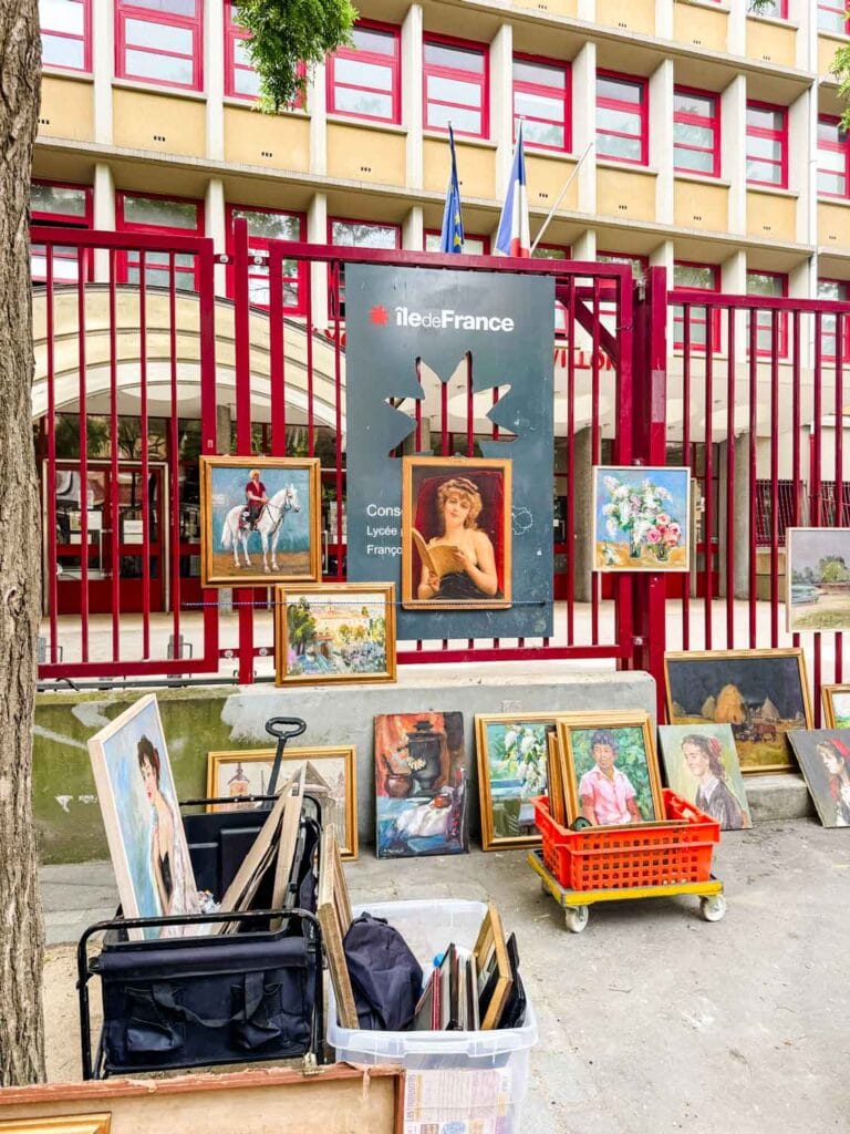 Vintage artwork hangs on the back fence that separates the the sidewalk from the sports complex at the Vanves flea market in Paris.