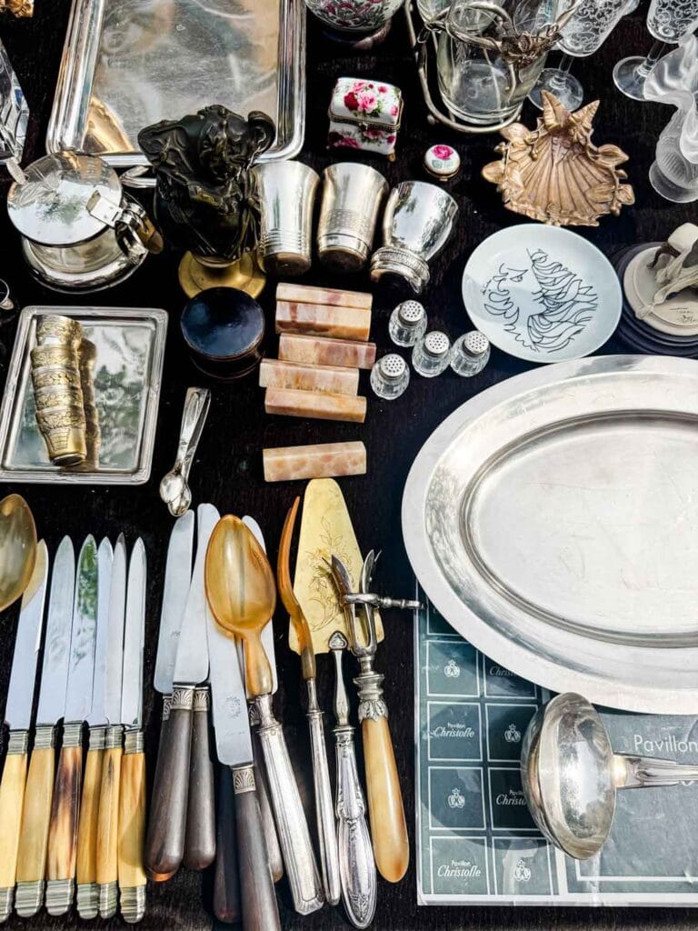 Vintage silverware, a large silver tray, and other vintage products are lying on this vendor's table at the Vanvas flea market in Paris.