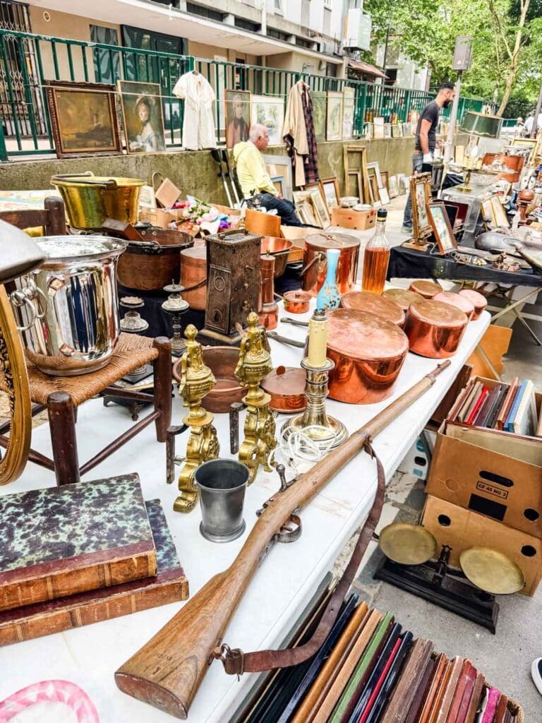 Copper pots, a vintage rifle, a silver wine bucket and other products are display on this table at a flea market in Paris.