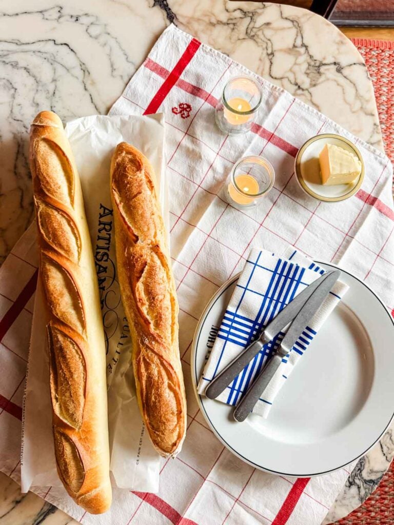 Two different types of baguettes are lined up on a vintage French tea towel next to plates, knives, butter, and little glass jars with votives.