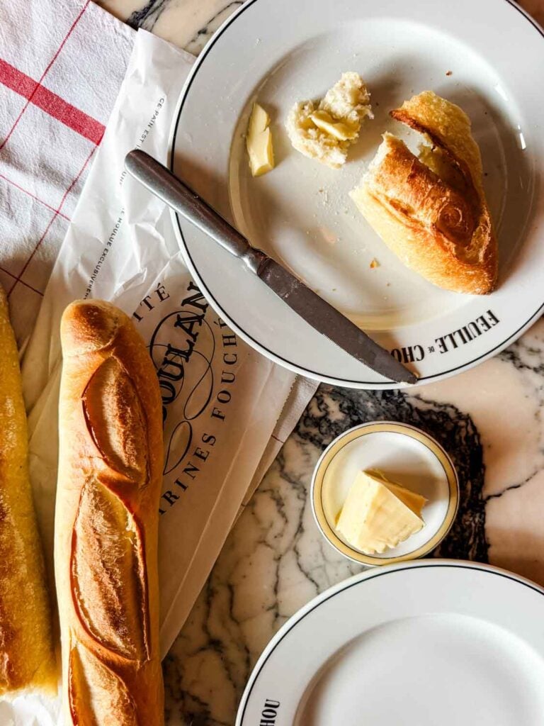 Two baguettes are next to each on a table and pieces have been torn off and placed on a plate next to some French butter.