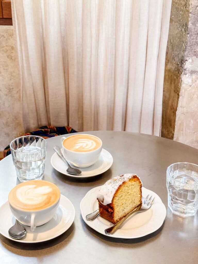 Coffee in cups, with water glasses, cake on a white plate with two forks on round table.