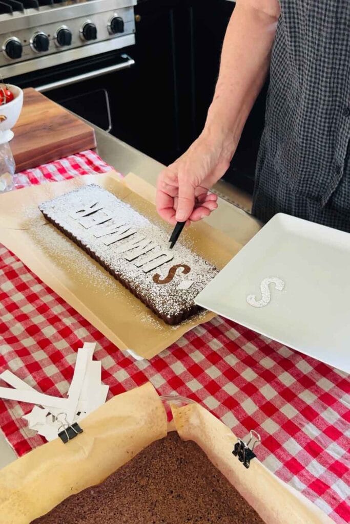 The letters on a brownie have  been dusted with powdered sugar and a woman is removing the letters with a pair of small tweezers.
