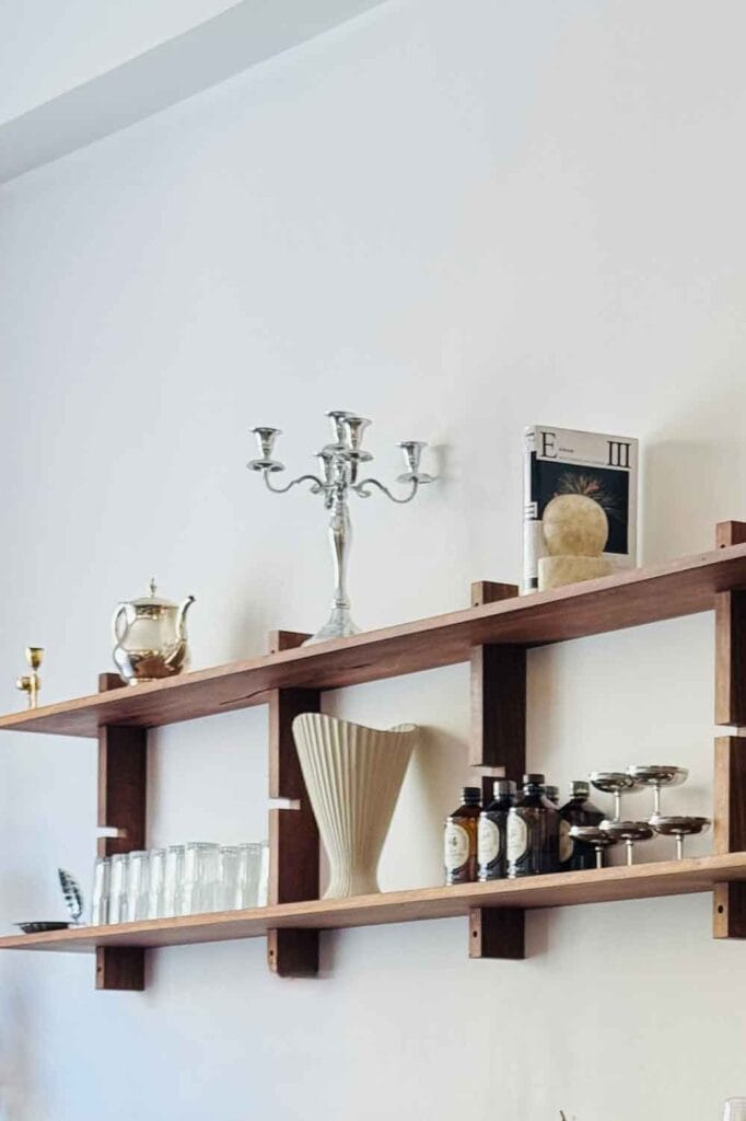Modern wall shelf with cadelabra, glasses, silver coffee pot, book, bottles.