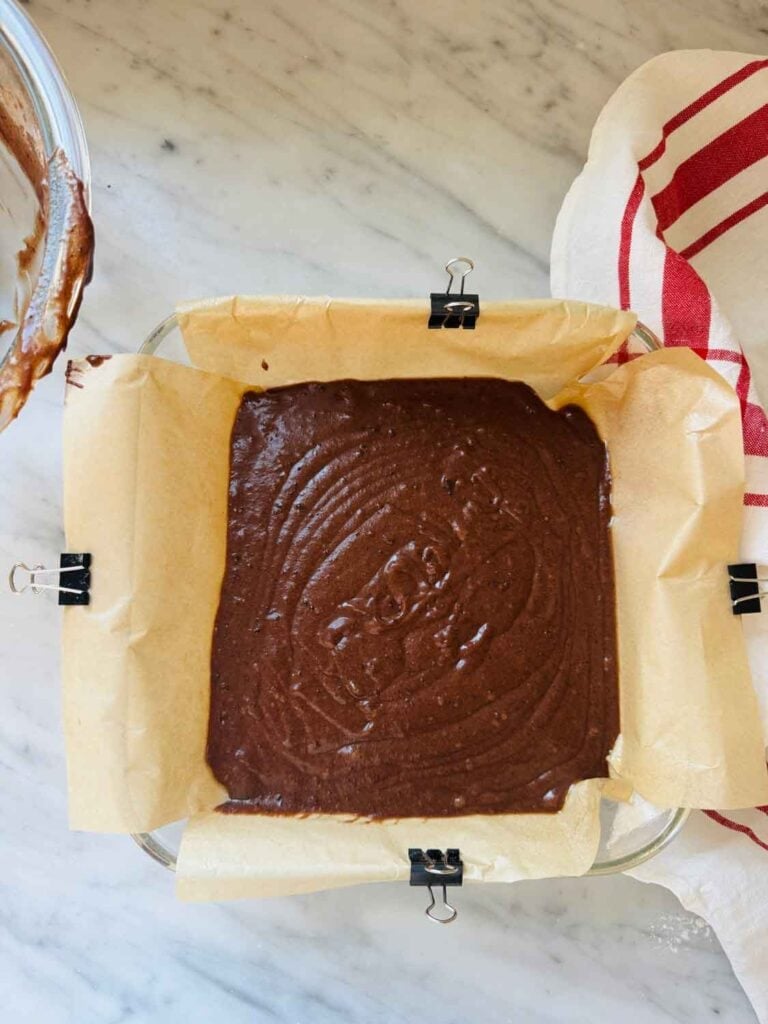 Brownie batter has been poured into and 8-inch cake pan lined with parchment paper held in place with small clips.