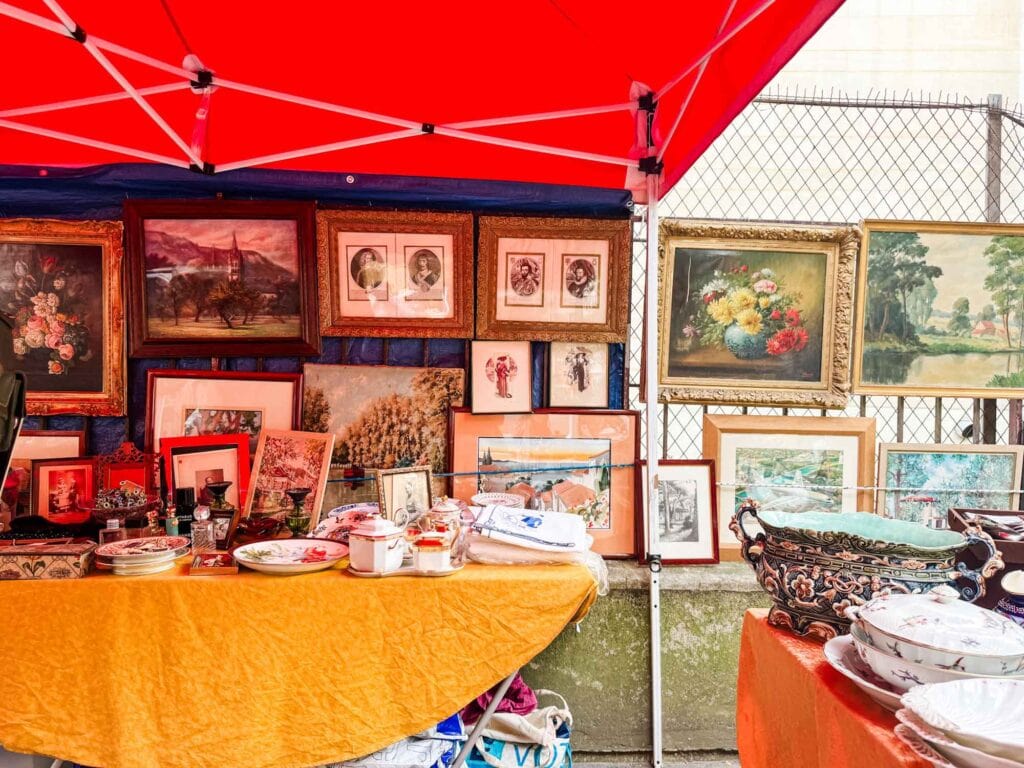 A red awning covers a stall at the Vanves Flea Market in Paris. Paintings are hanging on the wire fence behind it.