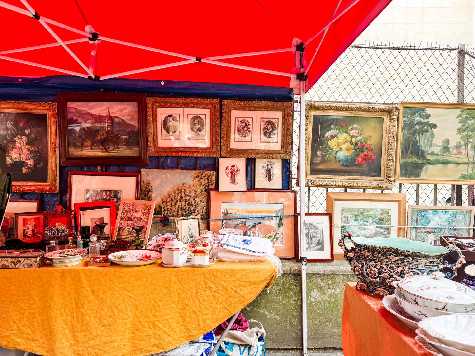 A red awning covers a stall at the Vanves Flea Market in Paris. Paintings are hanging on the wire fence behind it.