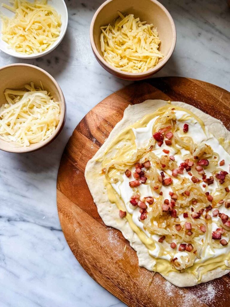 Pizza with bacon on peel, plus small bowls of shredded cheese.