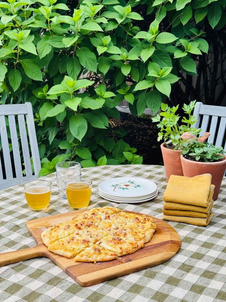 Garden setting with hydrangea tree in background, table with green checked cloth, glasses, plates, terracota panter, pizza on peel, gold napkins stacked.