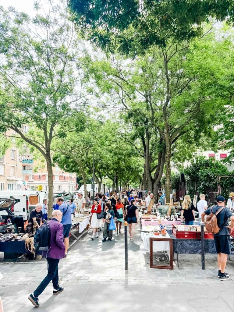 Crowds of people are walking down the tree-covered sidewalks of Portes Vanves Flea Market in Paris.