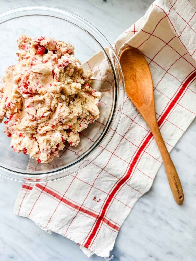 Bowl with cookie dough and freeze dried strawberries in it the dough, next to a red and cream dish towel and a wooden spoon. 