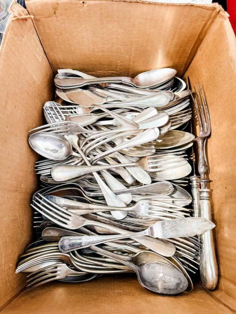 Vintage forks, knives, and spoons are piled into a cardboard box and sitting on a table.