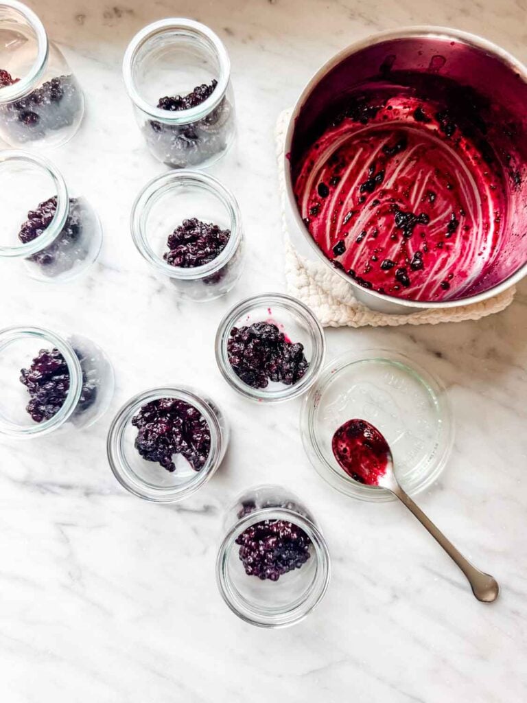 Glass jars are filled with blueberry compote and the empty saucepan is sitting next to the containers.