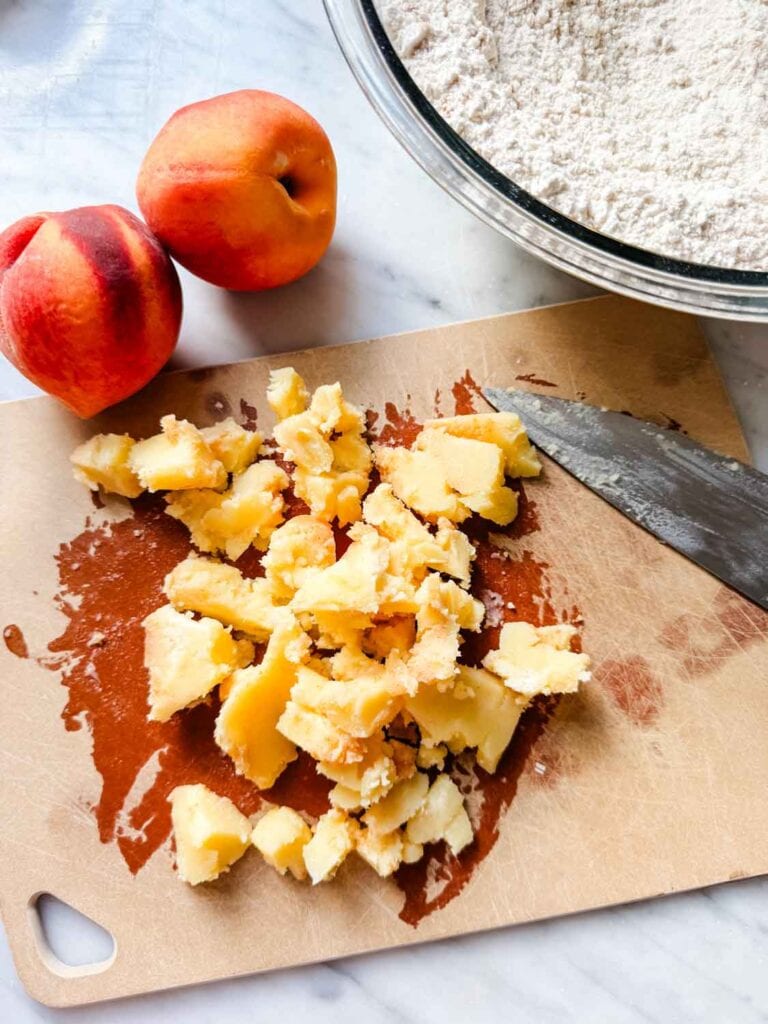 Cubed brown butter on a cutting board next to two ripe peaches and a bowl of the flour mixture.