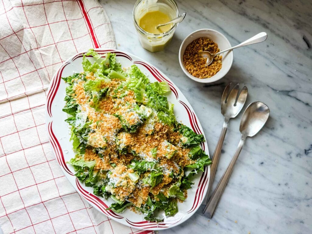 Caesar salad on platter with a red design on the rim, jar of salad dressing, bowl of panko breadcrumbs, and silver salad servers.