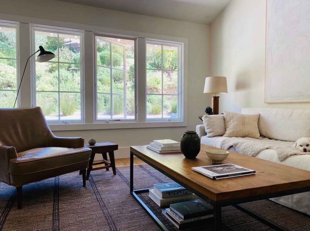 Modern living room with wood coffee table, sofa, chair, brown rug, big windows.