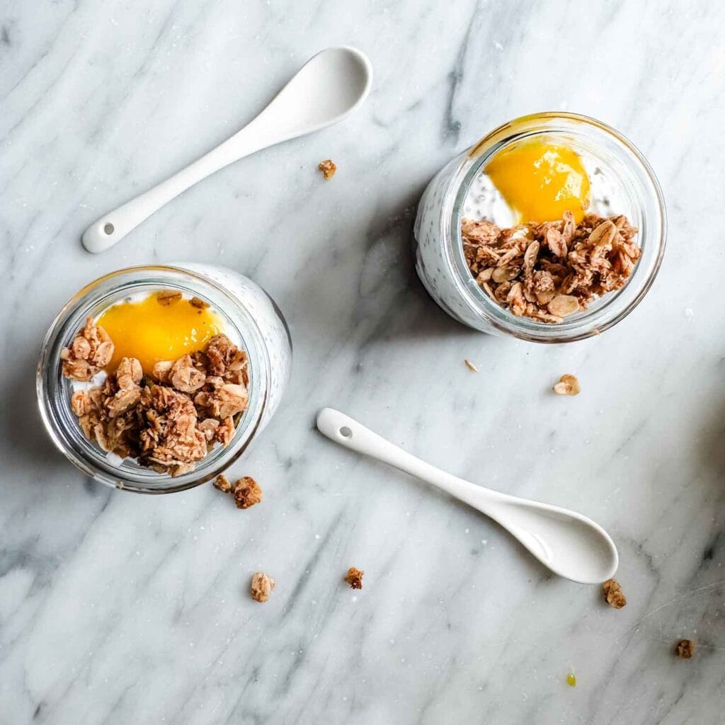 Two white porcelain spoons are sitting next to chia seed pudding that is topped with lemon curd and granola.