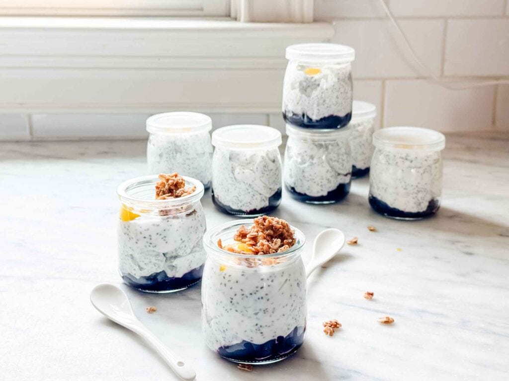 Containers of blueberry compote with chia pudding and granola topping are sitting an a kitchen counter. Small white spoons are sitting next to the jars.