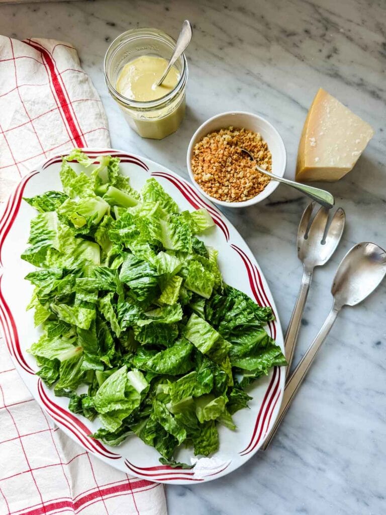 Caesar salad on platter with a red design on the rim, jar of salad dressing, bowl of panko breadcrumbs, parmsean cheese, and salad tongs.