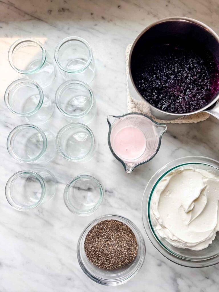 A saucepan with blueberry compote is sitting next to a measuring cup of Kefir, Greek yogurt, chia seeds, and empty glass jars.