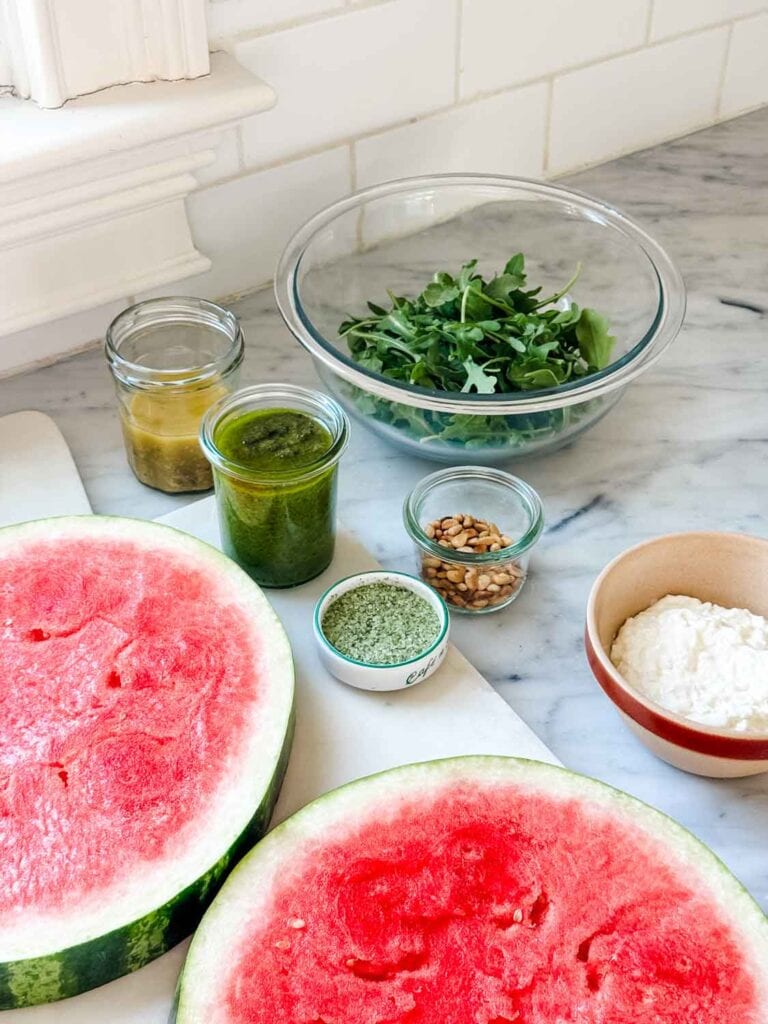 Watermelon slices are on a cutting board next to a small jar of pesto, vinaigrette, pine nets, stracciatella, and a bowl of arugula.