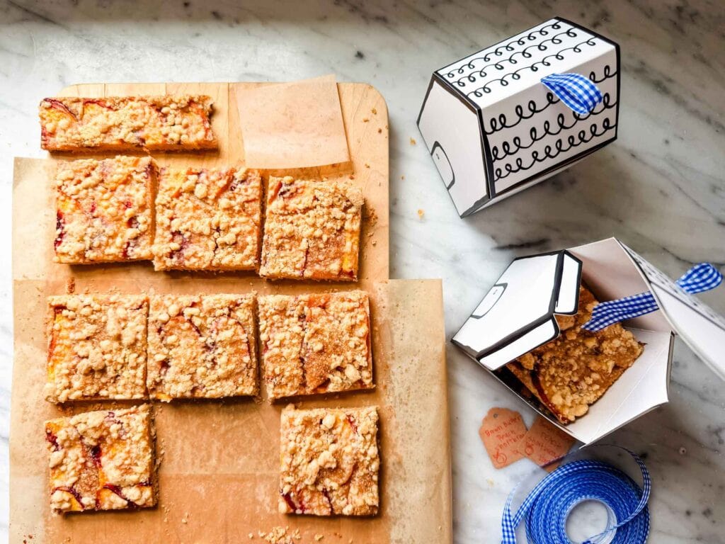 Brown butter peach shortbread bars are on a wooden cutting board next to house-shaped boxes with blue gingham ribbon that are filled with the peach boars.