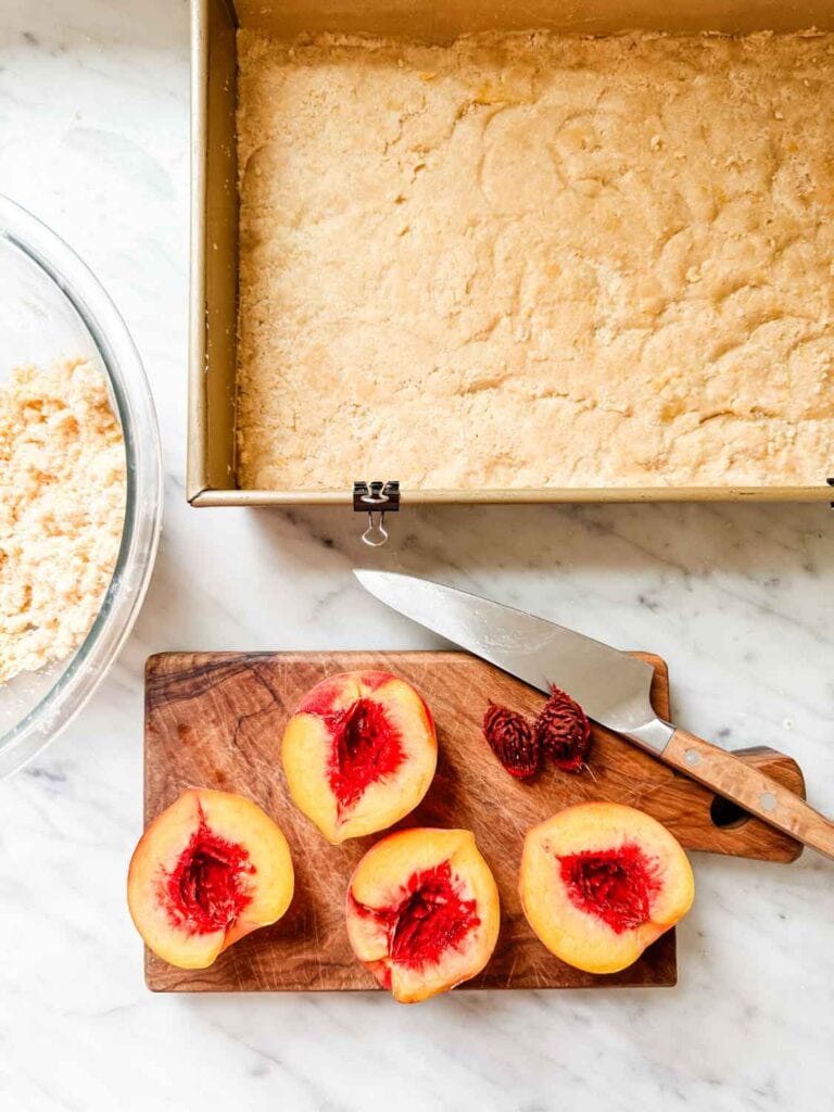 Brown butter shortbread crust is in a baking pan next to halved and pitted peaches.