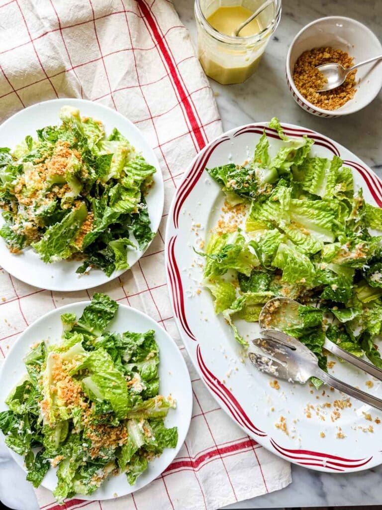 Caesar salad on platter with a red design on the rim, jar of salad dressing, bowl of panko breadcrumbs, and two plates with salad on them.