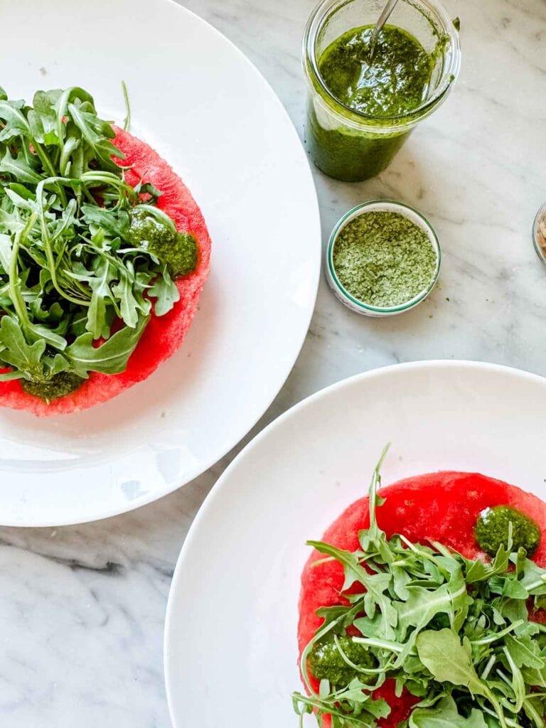 Watermelon rounds are served on two white plates and are topped with a handful of arugula. Pesto is the next ingredient.