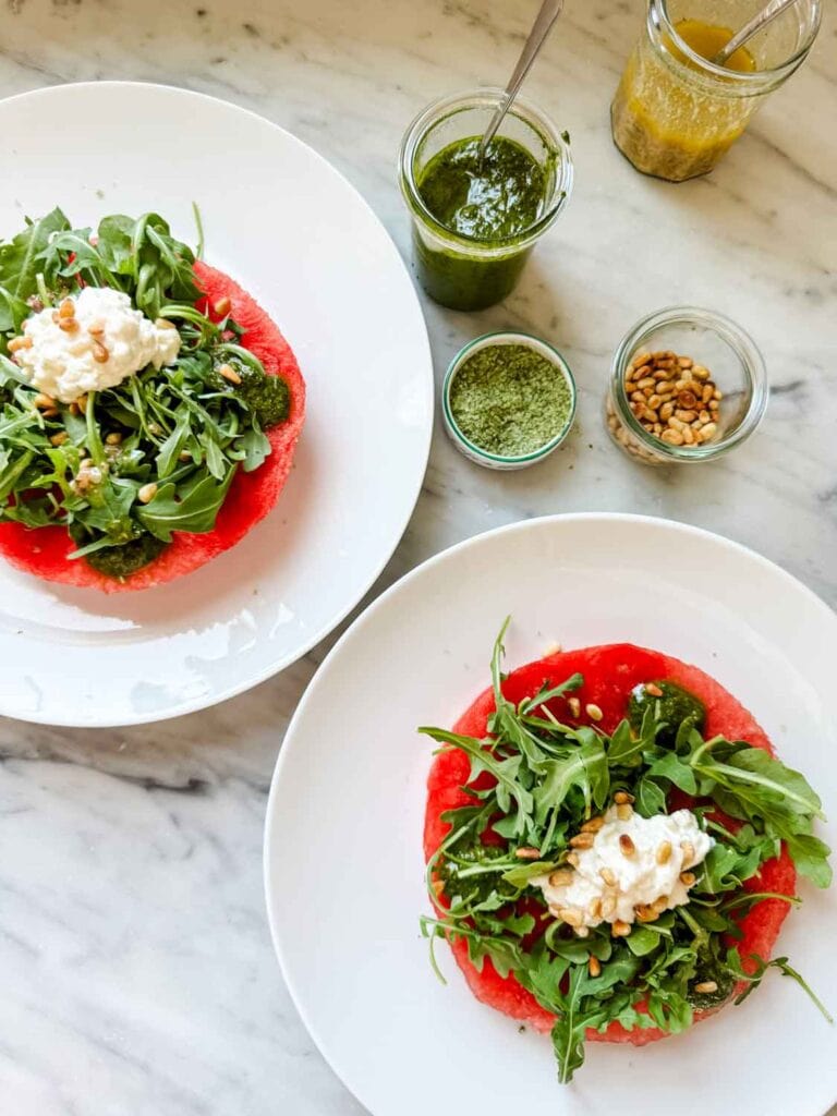 Two watermelon rounds on small white plates are served topped with arugula, pesto, vinaigrette, stracciatella and pine nuts.