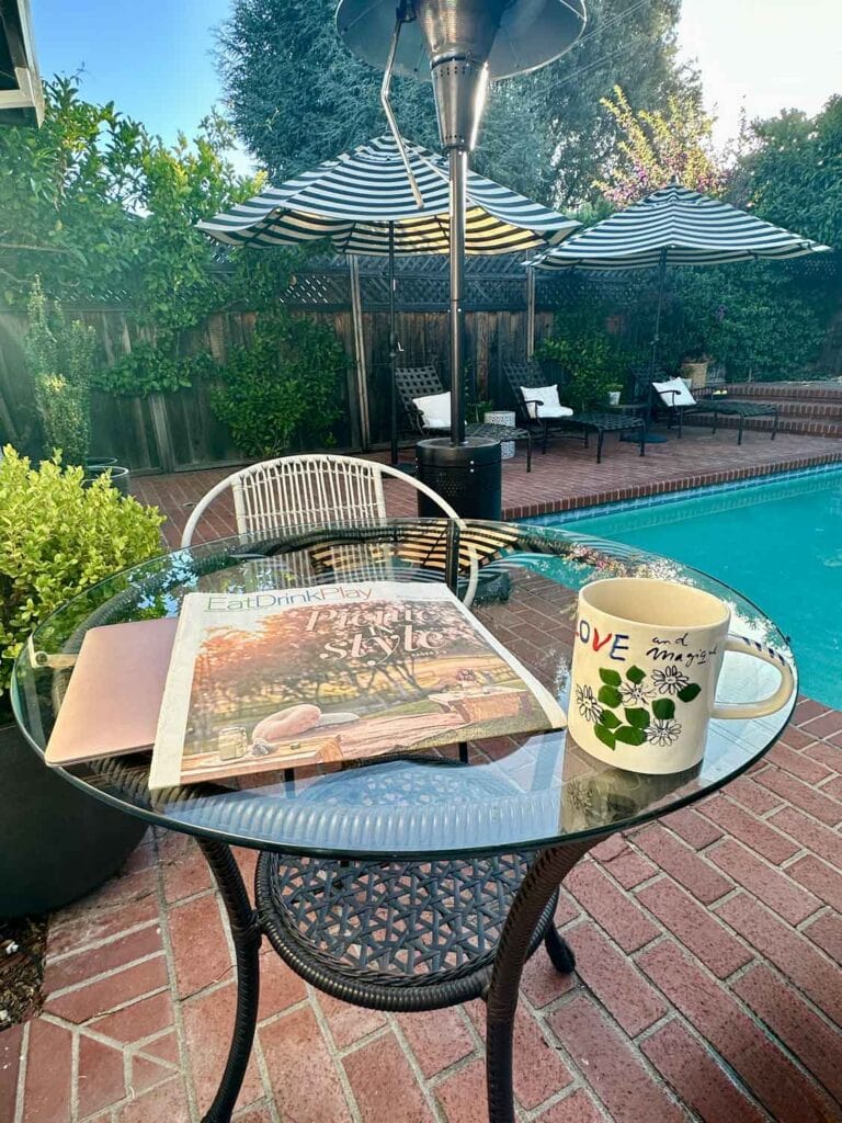 Small bistro table with magazine, mug by a pool with strioed patio umbrellas.