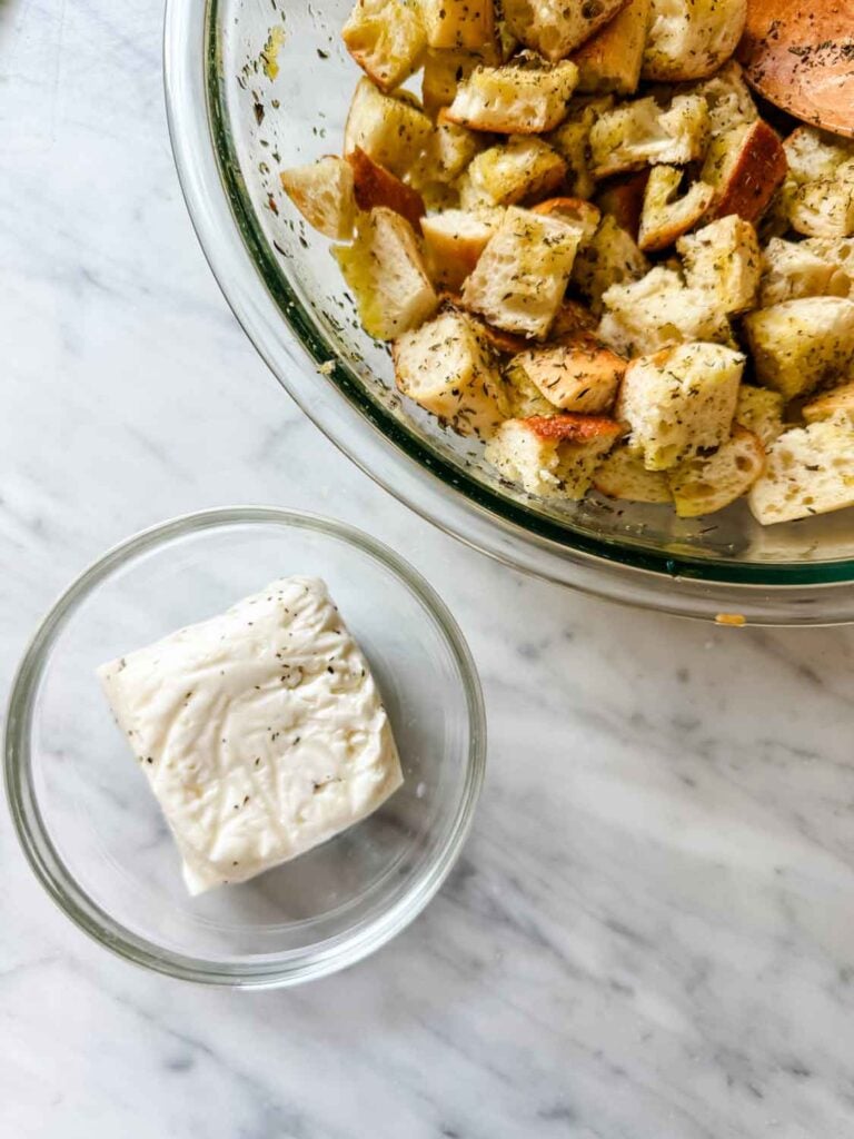 Cubed baguette dressed in dried herbs and Halloumi in a small glass bowl are prepared and ready for making croutons.