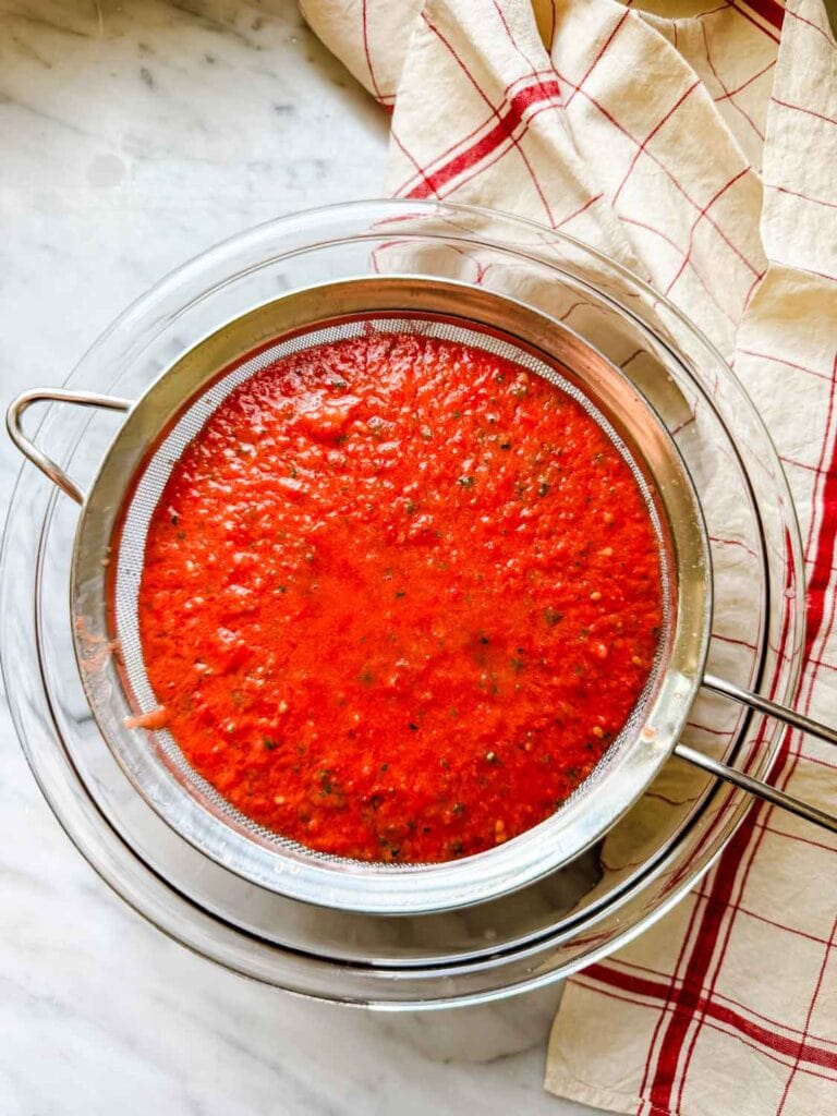Mesh strainer with chinckt gazpacho soup over bowl.