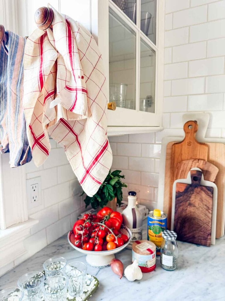 Tomatoes, salt, olive oil, small bottle of herbes de Provence, vinegar, shallot, garlic, basil on couter with cutting boards, and red plaid dish towel hanging.
