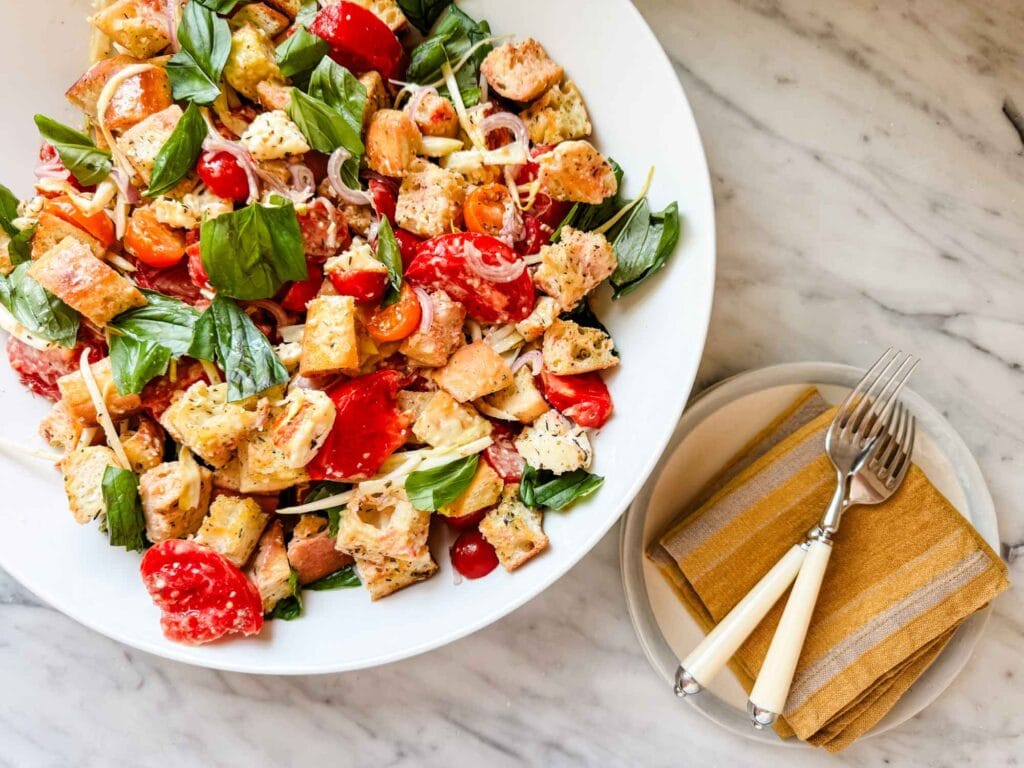 Panzanella salad with homemade Caesar dressing is tossed in a large white serving bowl sitting next to two small plates, salad forks, and cloth napkins.