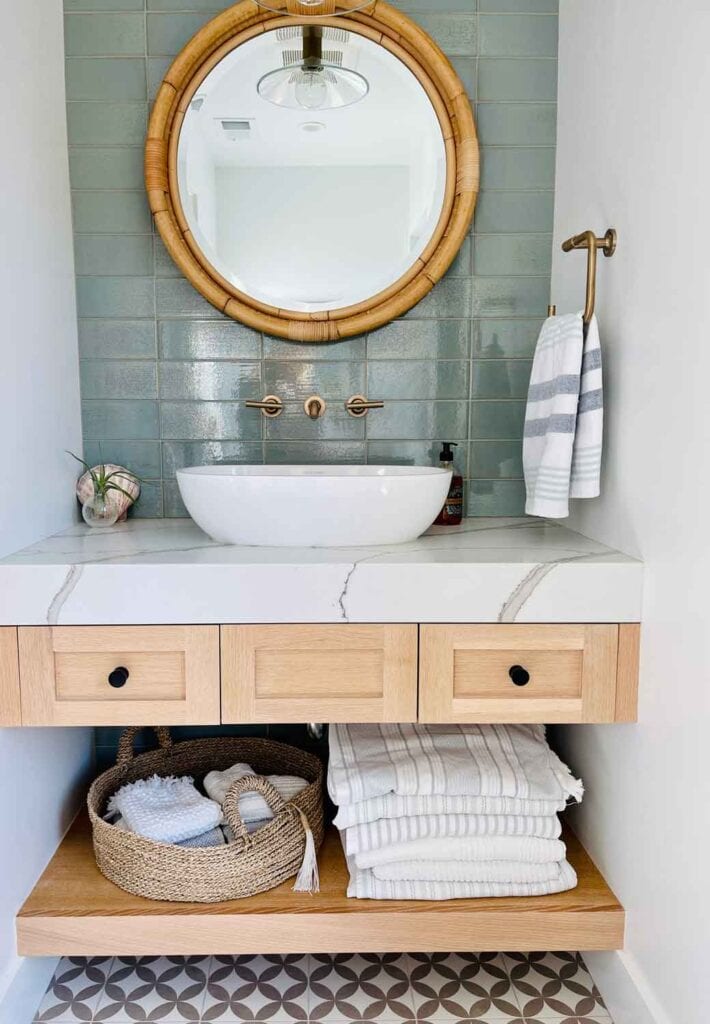 Round rattan mirror above white sink, vanity with stacks of towels, and a basket.