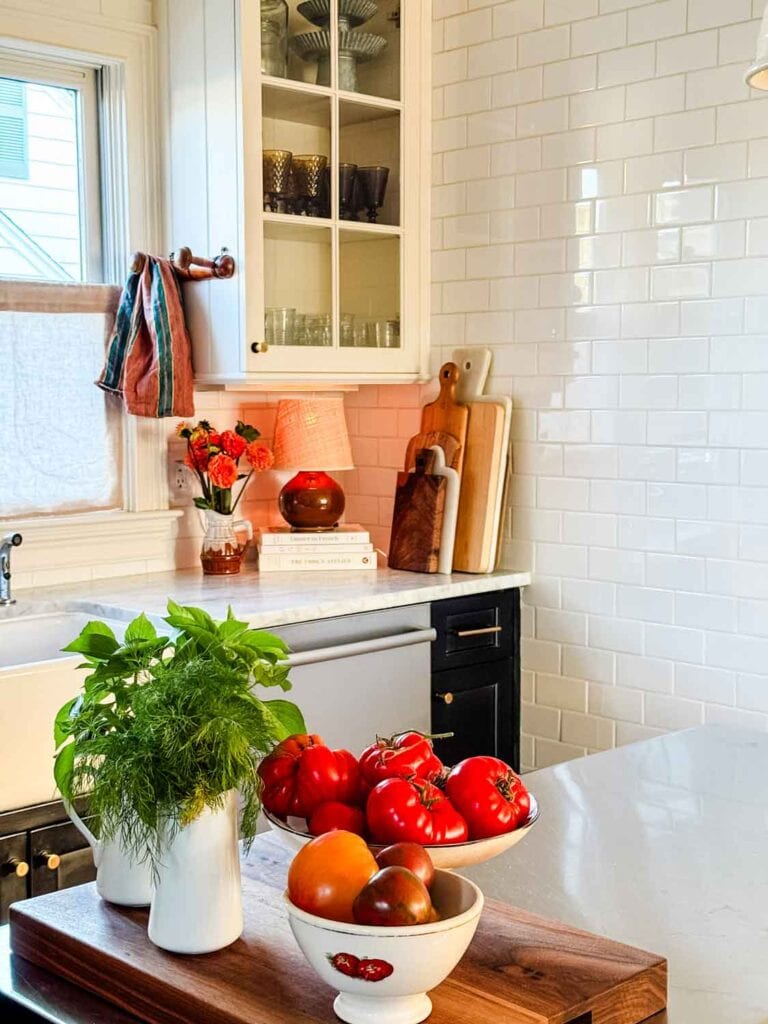 Corner of a kitchem bowl of tomatoes, small lmap on stack of cookbooks, flowers in pitcher.