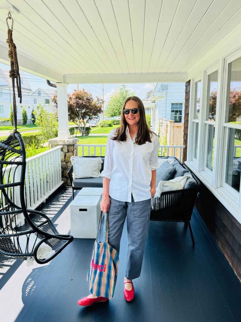 Woman wearing gray pants, white shirt, red ballet flats on a porch, holding a plaid tote bag.