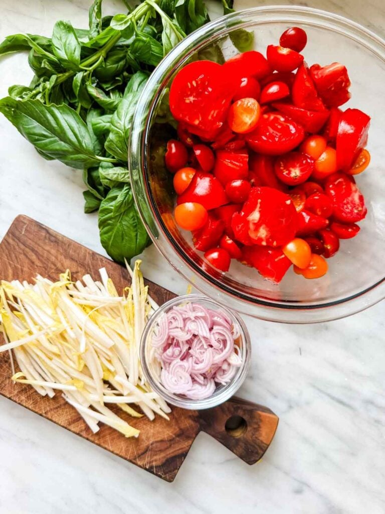 Chopped fresh tomatoes are in a glass bowl next to fresh basil, julienned endives, and sliced shallots for making Panzanella salad with Caesar dressing.