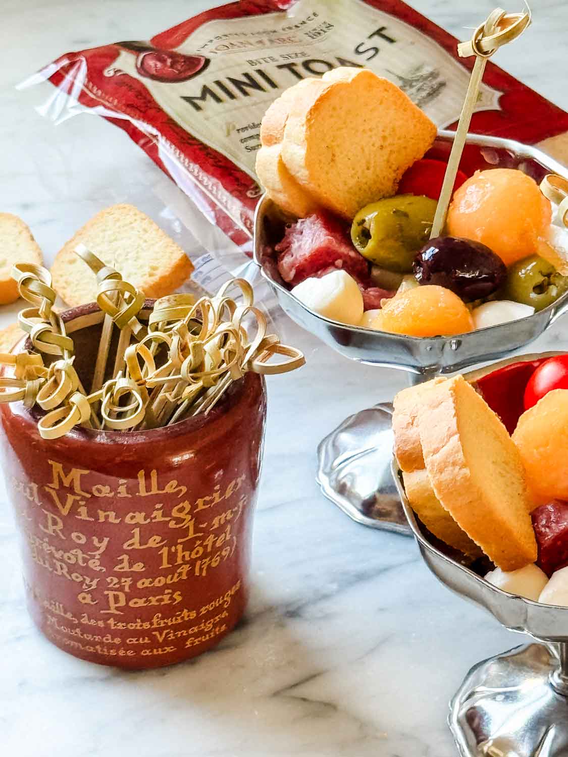 Charcuterie in small stainless dishes with package of mini toasts, and vintage musstard jar with picks.