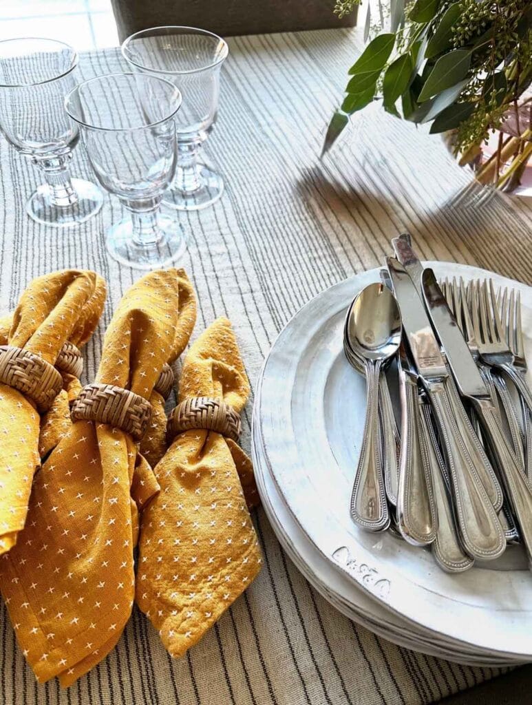Gold woven napkins with rattan napkin rings, stack of cermaic plates, silverware, glasses.