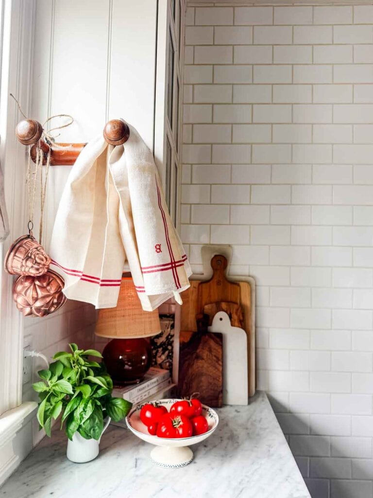 Pichter of basil, copper molds hanging from a vintage peg rack, tea towel, and a footed bowl with tomatoes. 