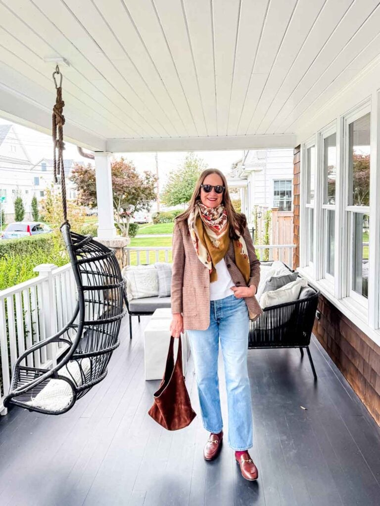 A women, wearing a scarf and fall blazer is on the porch of a home.