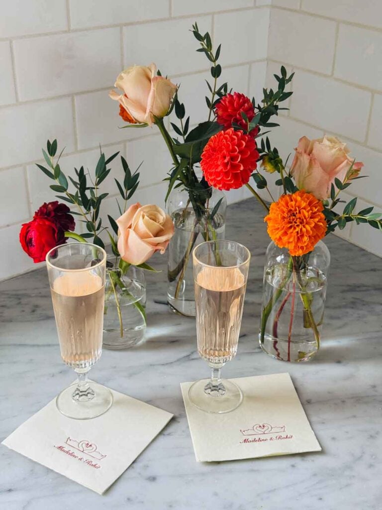 Champagne in flutes sit on customized napkins are next to small vases of colorful fall flowers.