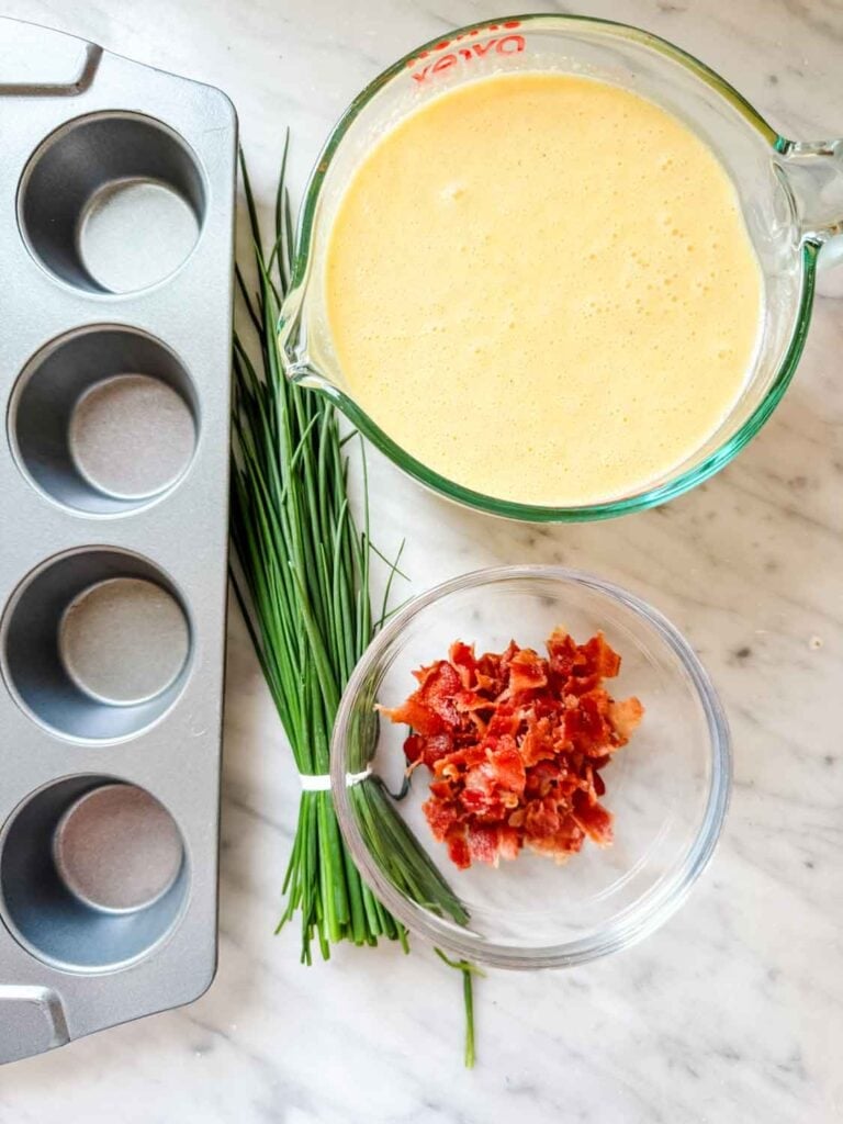 Cottage cheese egg mixture is in a glass measuring cup and sitting next to cooked bacon and a bunch of fresh chives.