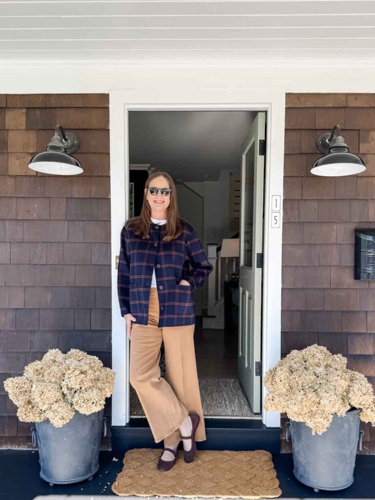 Annie Diamond wearing a navy sweater coat and denim trousers in rustic teak color in a doorway with dried hydrangeas.